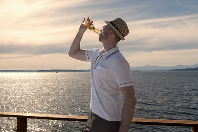 Man drinking beer by sea while wearing hat against sky