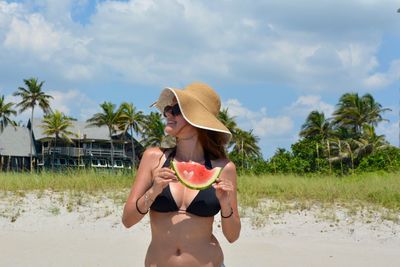 Young woman wearing bikini holding watermelon while standing at beach against cloudy sky