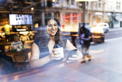 Happy young woman sitting with coffee cup and using mobile phone in cafe