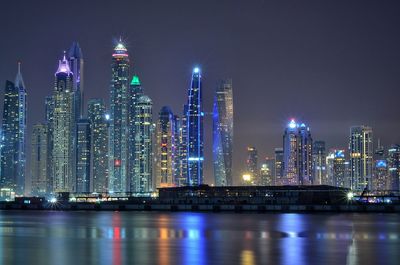 Illuminated modern buildings by river against sky at night