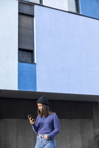 Young woman using phone while standing against building