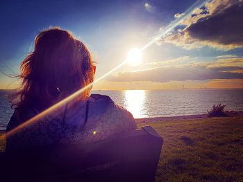 Rear view of woman sitting by sea against sky during sunset