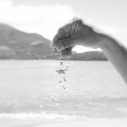 Close-up of hand pouring sand grains with  water background against sky