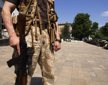 Midsection of military man holding rifle while standing in city