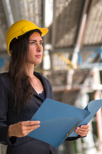 Absorbed engineer wearing a protective hardhat looking upwards and holding her project plan