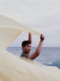 Young man holding scarf while standing at beach against sky
