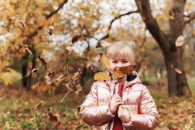 Happy little blonde girl plays with yellow autumn leaves in garden. child smiles, has fun in park