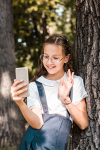 A schoolgirl in glasses makes a video call on a smartphone near a tree in the park. vertical view
