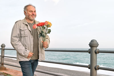 Happy smiling mature bearded man with bouquet of flowers walking towards his girlfriend on seafront.