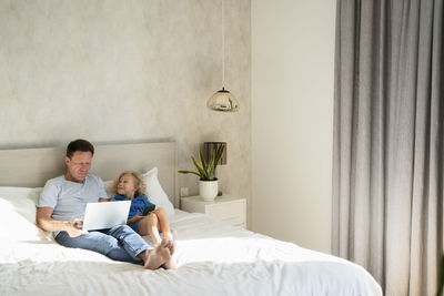 Smiling daughter with mobile phone by father using laptop on bed