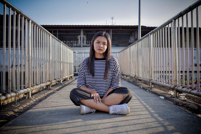 Full length portrait of teenage girl sitting on footbridge