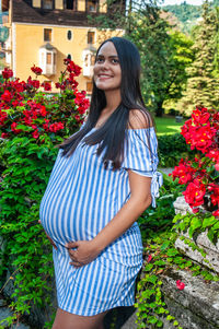Smiling beautiful pregnant south american girl, amidst flowers, wearing a long striped shirt
