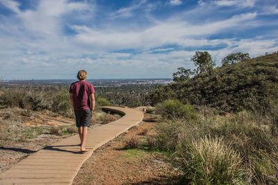 Rear view of mid adult man walking on boardwalk by plants against cloudy sky