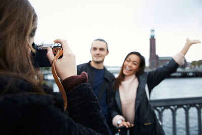 Woman photographing happy friends standing on bridge in city