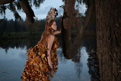 Beautiful young woman wearing headdress and leaves while standing by lake and tree trunk