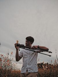 Young man holding tripod against sky