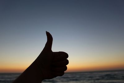 Silhouette cropped hand gesturing thumbs up on shore during sunset