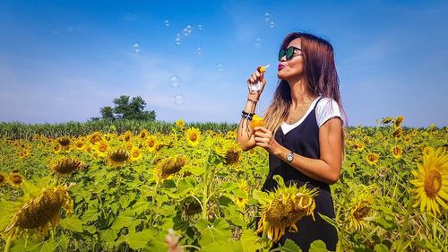 Woman blowing bubble while standing at farm against clear blue sky during sunny day
