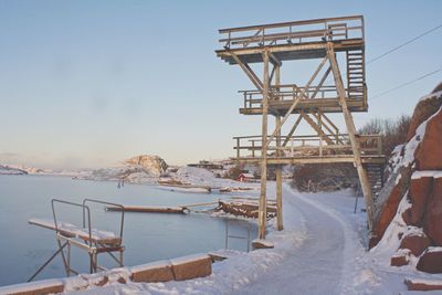 Wooden structure on snow covered land by sea against sky