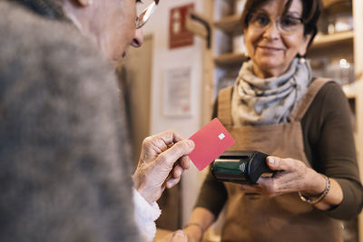 Side view of cropped unrecognizable positive female seller reaching out hand with terminal to happy senior woman doing payment with credit card in eco friendly store
