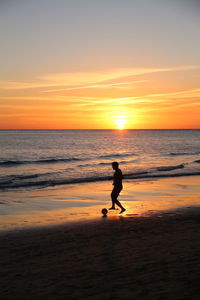 Full-length captured silhouette of a man playing football on the beach in front of sunset