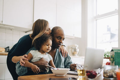 Woman embracing and kissing man with daughter using laptop at home