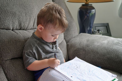 Young boy drawing doodles in a notebook while sitting on a couch at home