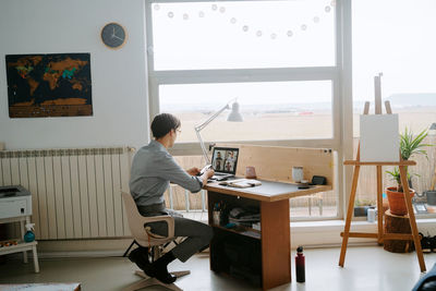 Side view of smiling male manager having video conference online via laptop while sitting at table and working from home during coronavirus epidemic