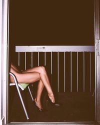 Low section of naked woman wearing high heels while sitting on chair in balcony seen through doorway at night