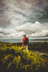 Rear view of man with backpack hiking on mountain against cloudy sky