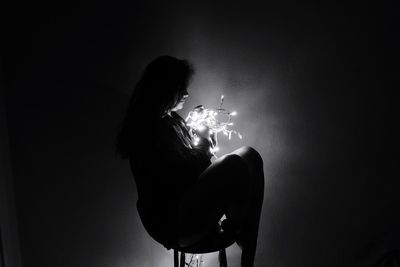 Side view of young woman holding illuminated light sitting on stool in darkroom at night