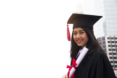 Portrait of smiling woman in graduation gown and mortarboard against buildings