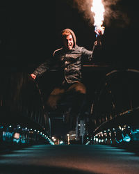 Portrait of man holding fire while jumping over road at night
