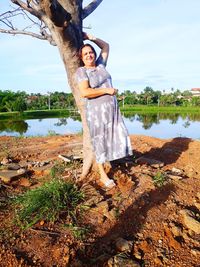 Portrait of woman leaning on tree trunk at lakeshore against sky