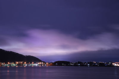 Illuminated city by sea against sky at night
