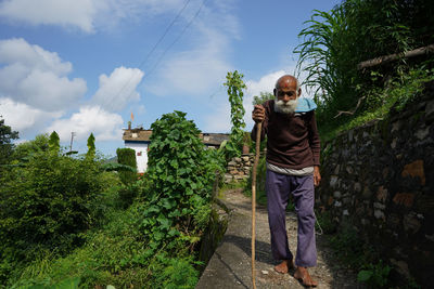 An old man with long white beard walking down a narrow pathway with the help of a stick.