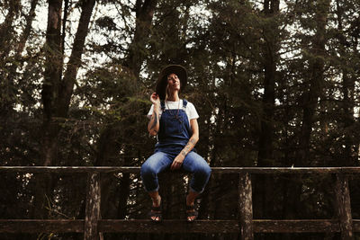 Full length of young woman sitting on railing in forest