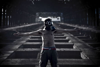 Shirtless man wearing gas mask with arms outstretched standing in abandoned building