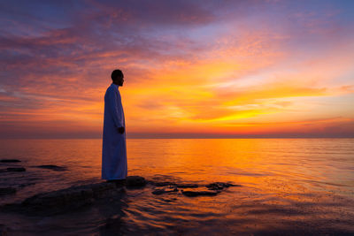 Rear view of woman standing at beach against sky during sunset