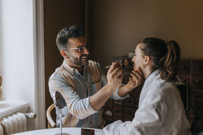 Smiling boyfriend applying make-up to non-binary person at home