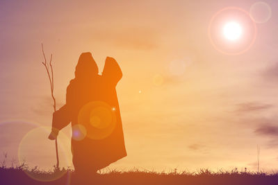 Silhouette person in halloween costume holding stick while standing on field against sky during sunset