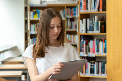 Smiling teenage girl holding file while standing in library