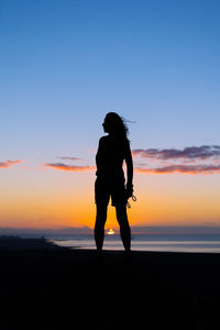 Side view of woman standing on beach against sky during sunset