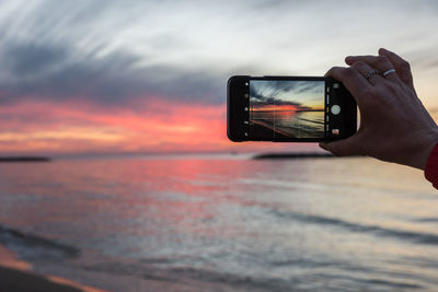 Cropped hand photographing sea with mobile phone during sunset