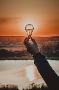 Cropped hand holding light bulb against sky during sunset