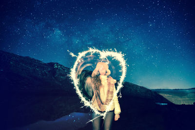 Woman making heart shape with sparklers against star field at night