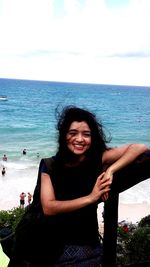 Portrait of smiling young woman standing at beach against sky