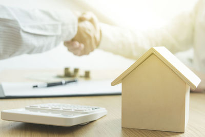 Close-up of model home and calculator on table against businessmen shaking hands in background