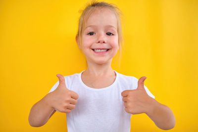 Happy joyful little girl smiling showing thumbs up isolated on yellow background