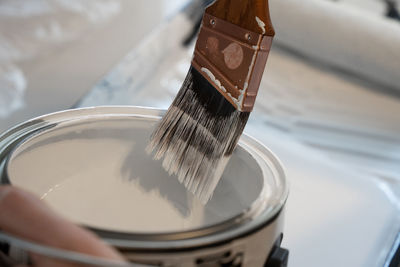 Paint brush is scraped on edge of paint can to remove excess white paint
